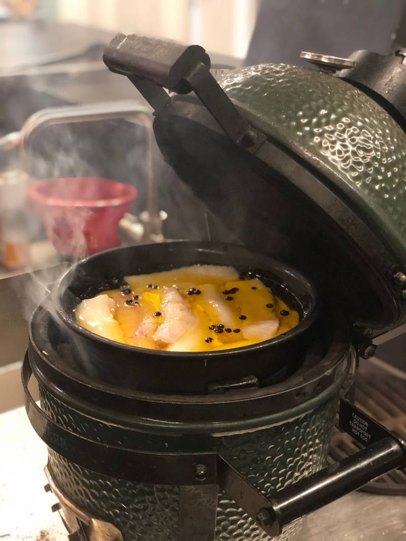 A pan of fish cooking in oil on a Big Green Egg, with the lid open and steam rising.